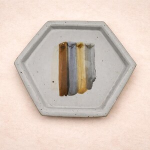 May include: A hexagonal, light gray ceramic tray with a rough texture. The tray holds five vertical brushstrokes of color: brown, blue-gray, yellow-gold, and gray. The background is a light pink.