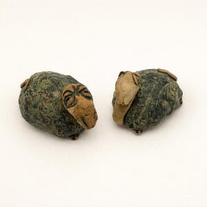 May include: Two ceramic sheep figurines. The sheep have a textured, mottled green body and tan-colored faces and ears. The figurines are placed on a white background.