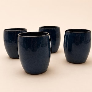 May include: Four navy blue ceramic tumblers with a speckled glaze. The cylindrical cups have a rounded base and a smooth, glossy finish. The cups are arranged on a light beige surface.