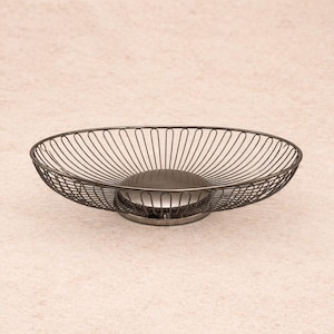 May include: A dark gray metal wire fruit basket with a circular base and a central, solid, dark gray disc. The basket has an oval shape with vertical wire supports. The design is modern and minimalist.