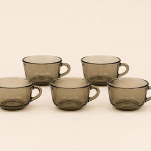 May include: A set of five matching tea cups made of translucent, smoke-gray glass. Each cup features a rounded bowl shape, a smooth rim, and a small, curved handle on the side. The glass has a uniform, dark gray tint throughout the entire structure.