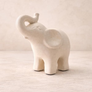 May include: A small, off-white ceramic elephant figurine with its trunk raised. The elephant has large ears and a textured surface, standing on four short legs. The figurine is a decorative item.