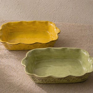 May include: Two rectangular ceramic baking dishes. One is a vibrant yellow, the other a soft green. Both have wavy edges and small handles. The dishes are on a neutral-toned surface, suggesting a rustic or farmhouse style.