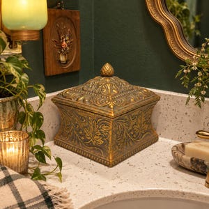 May include: A square, lidded box with a metallic gold and dark bronze finish, featuring intricate embossed leaf patterns and scrollwork. The lid is topped with a small, textured pinecone-shaped finial, and the base has a flared design.