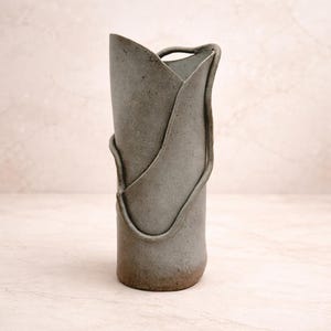 May include: A tall, gray ceramic vase with a unique, organic design. The vase features a cylindrical body with a folded, petal-like top and a decorative, looping accent. The base has a darker, earthy tone.