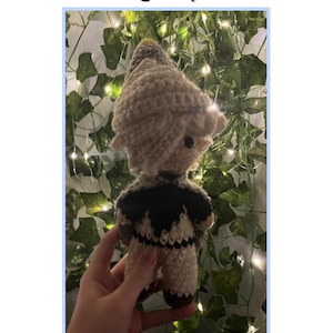 Puede incluir: Una muñeca de ganchillo con un gorro puntiagudo de color crema, pelo claro y una capa oscura sobre un conjunto de color crema. El texto Crochet Qifrey Pattern! aparece sobre la muñeca, que se muestra ante un fondo de hojas verdes.