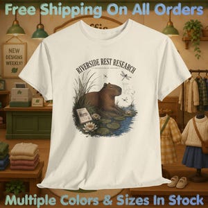 May include: A cream-colored short-sleeve t-shirt featuring a graphic illustration of a capybara sitting among water lilies and reeds. The text reads RIVERSIDE REST RESEARCH and UNHURRIED BY DESIGN. The design includes dragonflies and a small book.