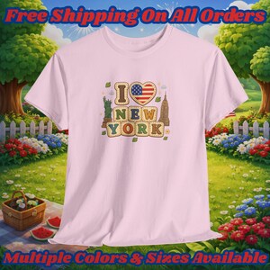 May include: A light pink t-shirt featuring a graphic print with the text I LOVE NEW YORK. The design includes a heart shaped American flag, an illustration of the Statue of Liberty, and the Empire State Building, surrounded by small leaf icons.