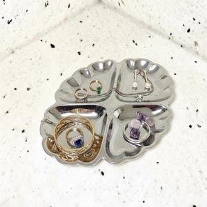 May include: A silver-toned, four-compartment metal jewelry tray with a scalloped edge. The tray holds various pieces of jewelry, including a blue gemstone ring, a gold-toned bangle, a green stone ring, pearl earrings, and purple stones.