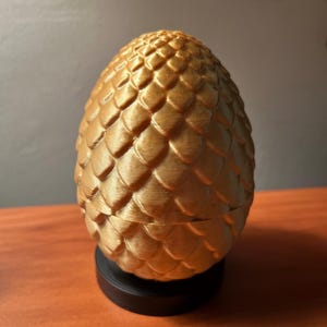 May include: A golden-colored, egg-shaped decorative item with a textured, scale-like pattern. The egg is resting on a black circular base, and the top half appears to be removable. The item is likely a decorative container or ornament.
