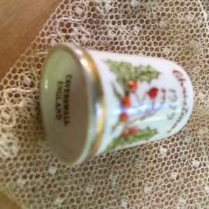 Puede incluir: Dedal de cerámica blanca con borde dorado y diseño de acebo verde. El dedal dice "Christmas 1973" y "Caverswall England".