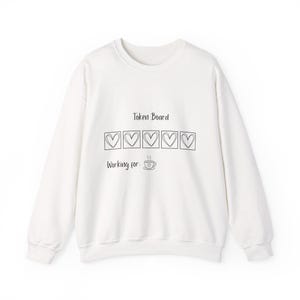 May include: Cream-coloured sweatshirt featuring the words "Token Board" above five heart-shaped boxes. Below, it reads "Working for:" and a coffee cup graphic. A comfortable, casual top for everyday wear.