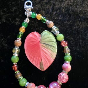 May include: A beaded keychain with a heart-shaped pendant. The keychain features a variety of colorful beads, including green, yellow, pink, and brown. The pendant is a leaf design with pink and green hues, and the keychain has a silver clasp.