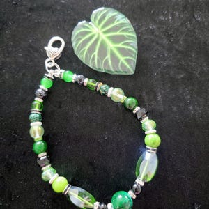 May include: A beaded bracelet with a variety of green beads, including round, oval, and faceted shapes, interspersed with black and silver accents. A silver heart-shaped clasp secures the bracelet. A green leaf-shaped decorative element is also present.