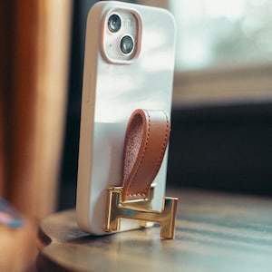 May include: A white smartphone case with a brown leather hand strap and a gold-colored metal stand. The phone case has two camera lenses and is resting on a wooden surface.