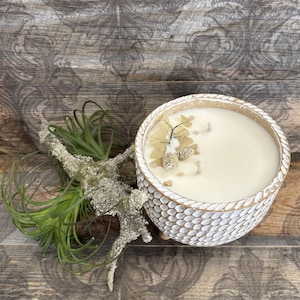 Puede incluir: Una vela redonda blanca en un cuenco texturizado con un diseño similar a una cuerda. La vela está adornada con pequeñas piñas y ramitas verdes. La vela está sobre una superficie de madera.