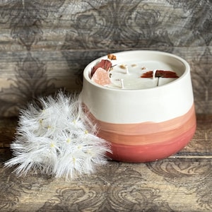 Può includere: Una candela in ceramica bianca con una sfumatura di pesca e corallo. La candela è decorata con cristalli e fiori secchi. Una pianta bianca e soffice è accanto alla candela. La candela è su una superficie di legno.