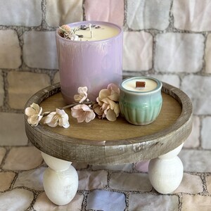 Peut inclure: Un petit plateau rond en bois avec trois pieds blancs en forme de boule, contenant deux bougies. L'une est dans un verre lavande, l'autre dans un petit récipient en céramique vert. Le plateau est décoré de fausses fleurs.