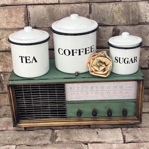 Pode incluir: Três potes de esmalte branco com as etiquetas "TEA", "COFFEE" e "SUGAR" em letras pretas. Eles estão em cima de um rádio verde vintage com acabamentos dourados. Uma flor decorativa é colocada entre os potes.