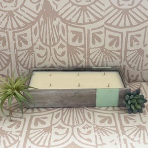 May include: A rectangular wooden candle holder with a cream-colored candle and multiple wicks. The holder has a distressed gray finish with a mint green accent. Decorative succulents and air plants are placed near the candle.