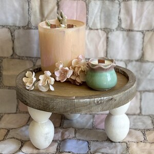 Puede incluir: Un alza decorativa de madera con una parte superior redonda y tres patas de bola blancas. Contiene una vela grande de color melocotón con una mecha de madera, una vela verde pequeña y flores rosas. El fondo es un patrón de ladrillos.