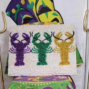 May include: A square canvas art piece featuring three beaded lobster designs in purple, green, and gold. The lobsters are set against a white background, surrounded by white beads. Two gold trident props flank the artwork, with a Mardi Gras theme.