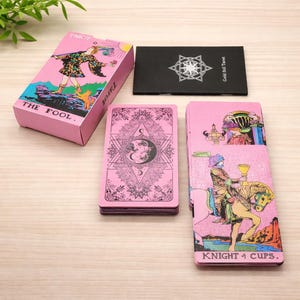 Puede incluir: Un juego de cartas de tarot que incluye una caja rosa con la etiqueta THE FOOL, un folleto negro titulado Gold foil Tarot, un mazo de cartas con diseño de luna geométrica rosa y una carta que muestra una figura a caballo llamada KNIGHT of CUPS.