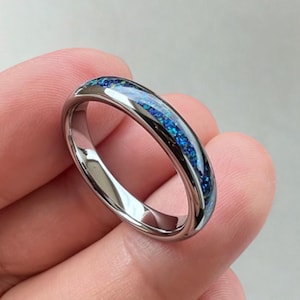 May include: A polished silver ring featuring a central inlay of blue and teal-coloured material. The ring's surface is highly reflective. The inlay has a speckled design, enhancing the ring's visual appeal.