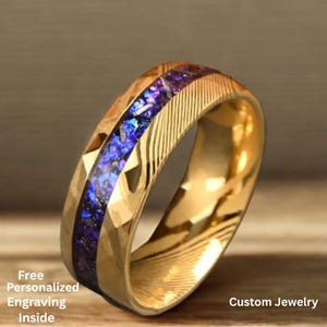 Könnte beinhalten: Ein goldfarbener Ring mit einem mittigen Einsatz aus schimmernden blauen und violetten Steinen. Der Ring hat ein strukturiertes Band mit geometrischen und linearen Mustern. Der Text "Custom Jewelry" ist sichtbar.