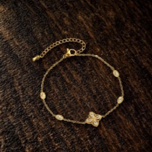 May include: Gold-toned bracelet featuring a delicate chain and a floral charm. The bracelet is adorned with oval-shaped, clear gemstones and a four-petal flower design. The adjustable chain measures approximately 7 inches.