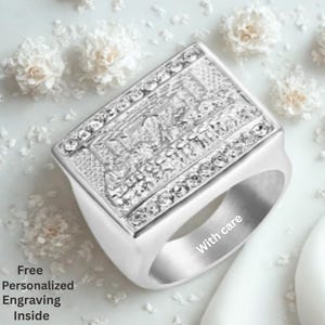 Può includere: Un anello sigillo rettangolare color argento con una raffigurazione in rilievo dell'Ultima Cena sulla parte frontale. La fascia è liscia e lucida, con il testo "With care" visibile sulla superficie interna. Il testo "Free Personalized Engraving Inside" è presente.