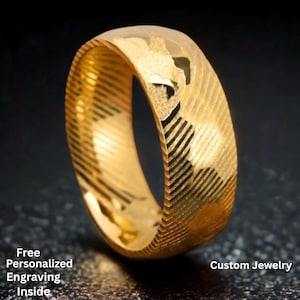 Puede incluir: Un anillo dorado con una superficie texturizada y líneas diagonales. El anillo tiene una banda ancha y un acabado pulido. El texto "Free Personalized Engraving Inside" y "Custom Jewelry" son visibles.