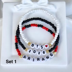 Könnte beinhalten: Set aus drei Armbändern mit Perlen. Ein Armband ist weiß, eines ist schwarz und eines ist eine Kombination aus roten und schwarzen Perlen. Die Armbänder haben weiße Buchstabenperlen mit der Aufschrift "BTS 2026 ARIRANG" mit goldenen Akzenten. Der Text "Set 1" befindet sich unten links.