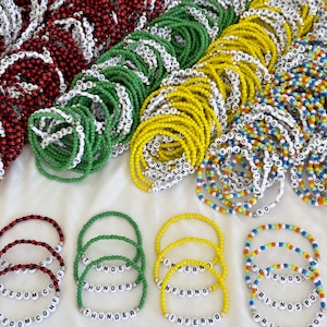 May include: A collection of handmade beaded bracelets in red, green, yellow, and multicolored patterns. Each bracelet features white letter beads spelling out words including Freedom, Thunder, Inferno, and Friends PDX, with small metallic accents.