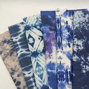 Può includere: Una collezione di sei campioni di tessuto piegati con vari motivi blu e bianchi. I design includono effetti tie-dye, forme geometriche a diamante, texture maculate e stampe astratte ispirate all'indaco disposte a ventaglio.