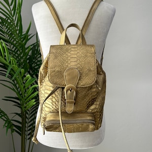 Puede incluir: Una mochila de piel de serpiente de color dorado con cierre de hebilla y correas ajustables.