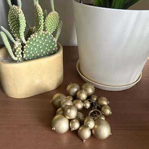 May include: A cluster of gold-coloured spherical beads of different sizes, linked by a chain. A small cactus in a yellow pot and a potted plant with green leaves are in the background.