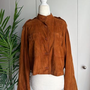 May include: A tobacco brown suede jacket featuring a high collar, a front button closure, and two chest pockets. The jacket has long sleeves with structured shoulders and epaulets, finished with a straight hemline that hits at the waist.