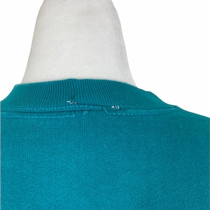 May include: Teal blue crew neck sweatshirt with a sewn seam detail on the back of the neck.