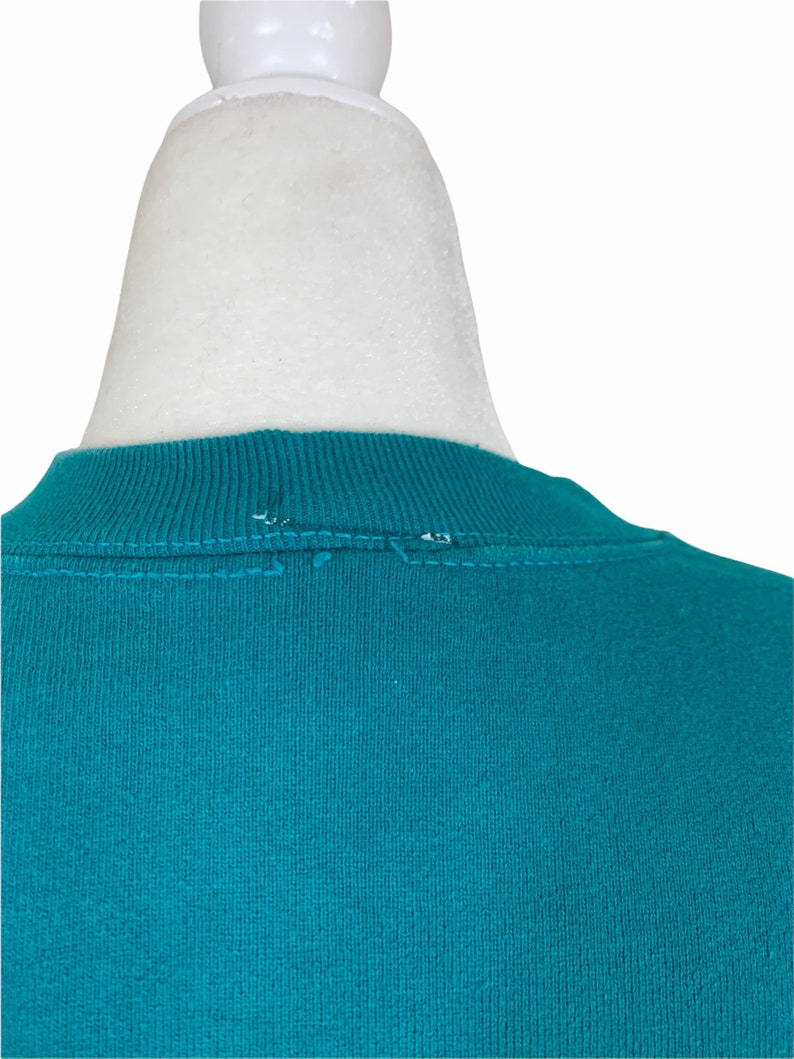 May include: Teal blue crew neck sweatshirt with a sewn seam detail on the back of the neck.