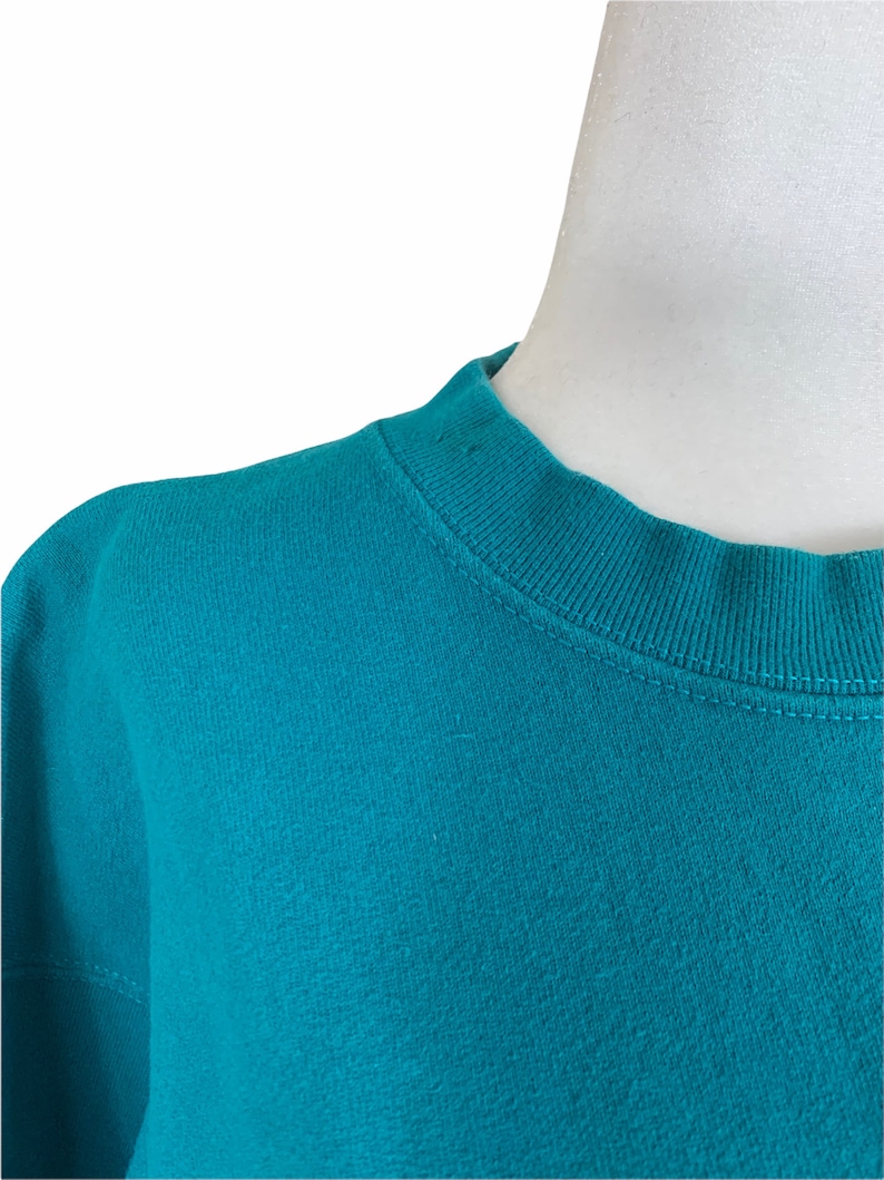 May include: Close-up of a teal blue sweatshirt with a ribbed crew neck.