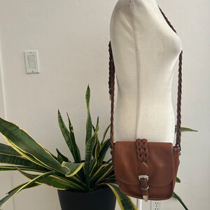 May include: A brown leather crossbody bag with a braided strap and a silver buckle. The bag is on a white mannequin.