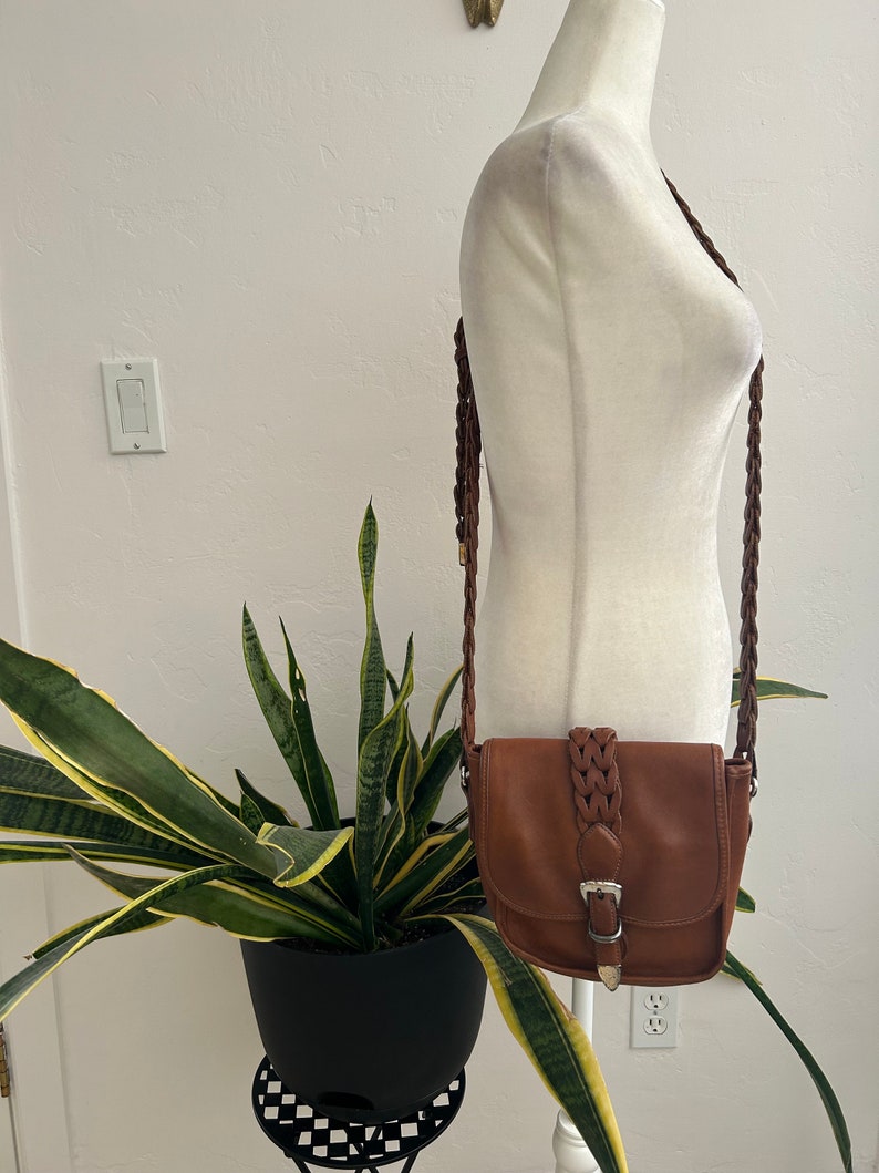 May include: A brown leather crossbody bag with a braided strap and a silver buckle. The bag is on a white mannequin.