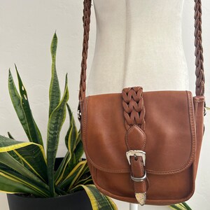 May include: A brown leather crossbody bag with a braided strap and a silver buckle. The bag has a flap closure and a small pocket on the front.