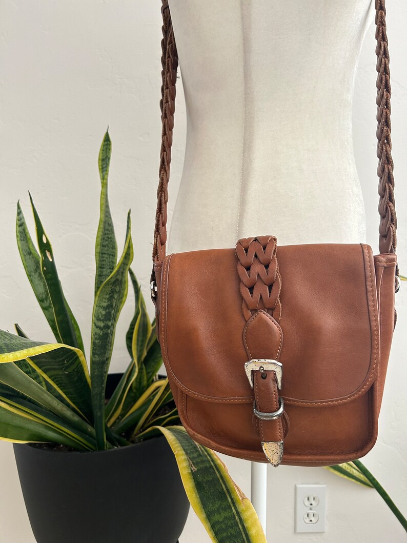 May include: A brown leather crossbody bag with a braided strap and a silver buckle. The bag has a flap closure and a small pocket on the front.