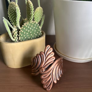 May include: A copper-coloured metal bracelet with a decorative design of overlapping leaves. The bracelet is on a wooden surface.