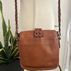 May include: A brown leather crossbody bag with a braided strap. The bag has a flap closure with a decorative braided strap detail.