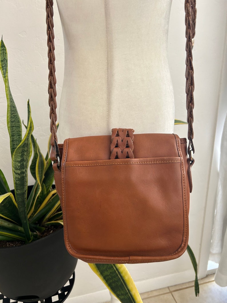 May include: A brown leather crossbody bag with a braided strap. The bag has a flap closure with a decorative braided strap detail.