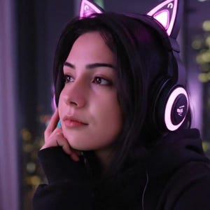 Op de afbeelding: Zwarte koptelefoon met kattenoren die oplichten met een roze gloed. De koptelefoon heeft een cirkelvormig ontwerp op de oorschelpen. De persoon op de foto draagt een zwarte hoodie.