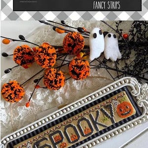 May include: A rectangular cross-stitch sampler featuring the word SPOOKY with orange jack-o'-lanterns, set in a white beaded frame. It is displayed with orange and black yarn pom-poms, white felt ghost shapes, and black decorative branches.